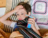 Chory chłopiec leży w łóżku, trzymając w dłoni inhalator. Jego ręka spoczywa na czole, a na szyi ma szalik. To mogą być objawy astmy.