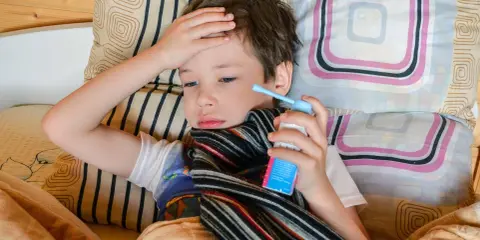 Chory chłopiec leży w łóżku, trzymając w dłoni inhalator. Jego ręka spoczywa na czole, a na szyi ma szalik. To mogą być objawy astmy.