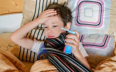 Chory chłopiec leży w łóżku, trzymając w dłoni inhalator. Jego ręka spoczywa na czole, a na szyi ma szalik. To mogą być objawy astmy.