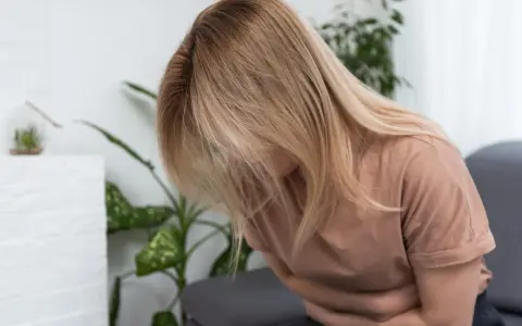Kobieta z blond włosami siedzi, trzymając się za brzuch. Wygląda na to, że cierpi z powodu zatrucia pokarmowego.