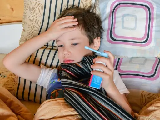 Chory chłopiec leży w łóżku, trzymając w dłoni inhalator. Jego ręka spoczywa na czole, a na szyi ma szalik. To mogą być objawy astmy.