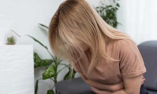 Kobieta z blond włosami siedzi, trzymając się za brzuch. Wygląda na to, że cierpi z powodu zatrucia pokarmowego.