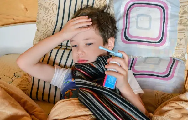 Chory chłopiec leży w łóżku, trzymając w dłoni inhalator. Jego ręka spoczywa na czole, a na szyi ma szalik. To mogą być objawy astmy.
